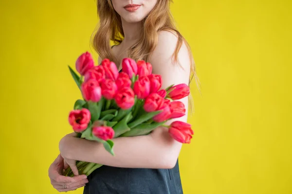 Eine unkenntliche Frau mit einem Strauß roter Tulpen auf gelbem Grund. Ein Mädchen in einem schwarzen Kleid hält einen Blumenstrauß in der Hand. Ein Geschenk zum Internationalen Frauentag. Frühjahrsferien. — Stockfoto