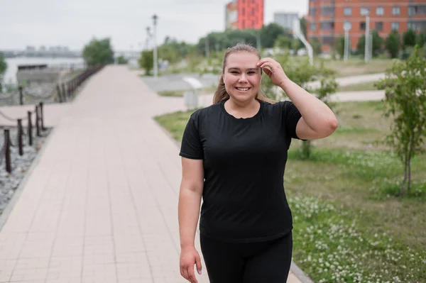 Eine übergewichtige junge Frau joggt im Freien. Dicke schöne lächelnde Mädchen in einem schwarzen Trainingsanzug ist in Fitness für die Gewichtsabnahme am Wasser engagiert. Eine Frau läuft an einem Sommertag. — Stockfoto