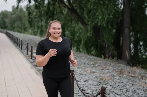 En fet ung kvinna som joggar utomhus. Fett vacker leende flicka i en svart träningsoverall är engagerad i fitness för viktminskning vid vattnet. En kvinna springer på en sommardag. — Stockfoto