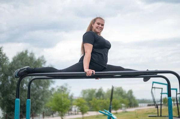 Eine schöne lächelnde, übergewichtige junge Frau streckt sich auf unebenen Stufenbarren im Freien. Dicke Turnerinnen beim Fitnesstraining auf dem Sportplatz. Sport draußen an einem warmen Sommertag. — Stockfoto