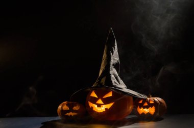 Üç ürkütücü Cadılar Bayramı balkabağı sislerin arasında karanlıkta parlıyor. Kara arka plan dumanlı bir cadı şapkası içinde kriko fener..