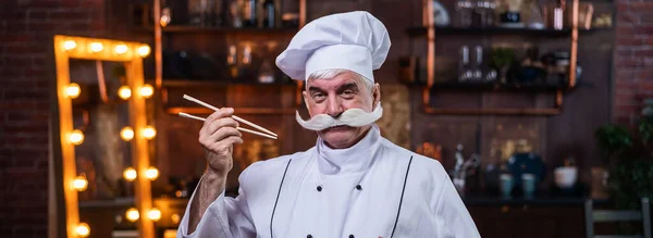 Um cozinheiro idoso amigável está segurando paus chineses. Um homem de cabelos grisalhos com um bigode cinza grosso trabalha na cozinha — Fotografia de Stock