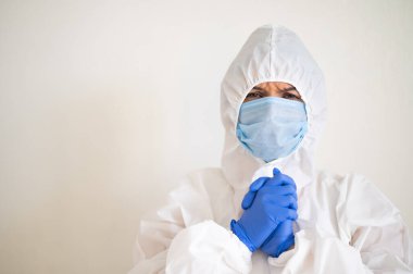 Koruyucu kıyafetli ve tıbbi maskeli bir kadın ellerini göğsüne koyup dua ediyor.