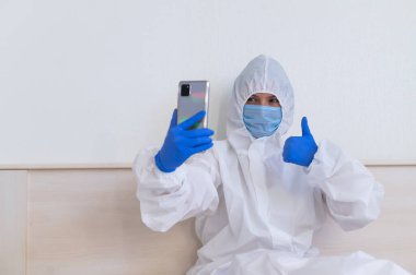 Koruyucu kıyafetli ve tıbbi maskeli bir kadın akıllı telefondan video bağlantısıyla iletişim kuruyor.