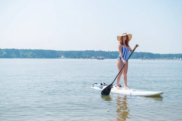 Una mujer en traje de baño y un sombrero posan en una mesa de sup en la playa. — Foto de Stock