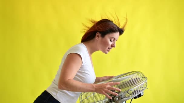 Egy gyönyörű vörös hajú nő lehűl, amint egy nagy elektromos ventilátor felett áll sárga háttérrel. Szőrös lány a szélben. Levegő hűtésére szolgáló készülék. — Stock videók
