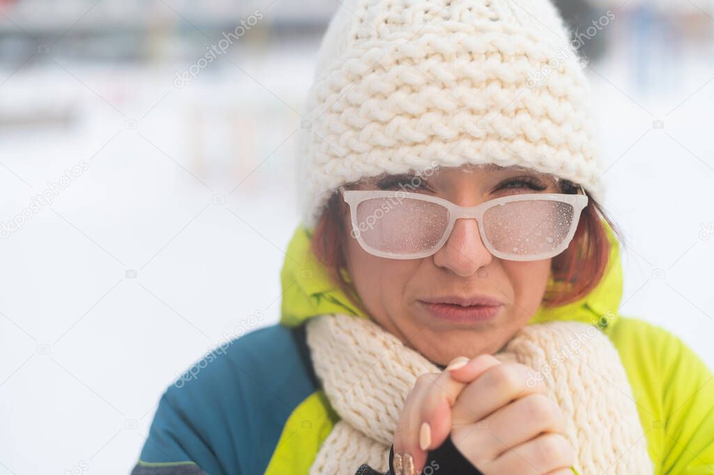 Portrait of a woman in glasses covered with hoarfrost. The girl is freezing and forgot gloves in very cold weather and blows on her bare hands