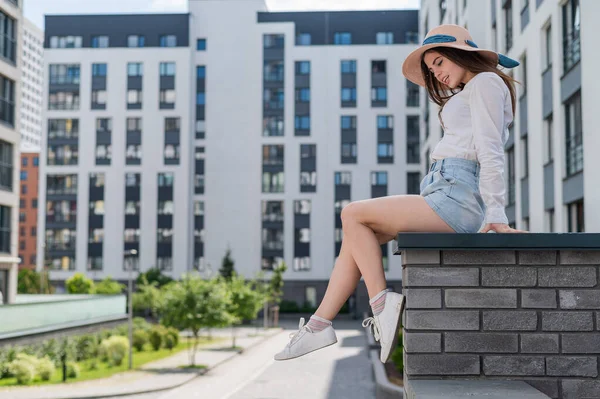 Hermosa joven en pantalones cortos y un sombrero posando mientras está sentada en un banco de piedra. Un retrato completo de una chica caucásica en un caluroso día de verano. —  Fotos de Stock