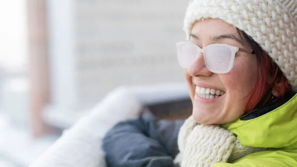 Χαμογελαστή λευκή γυναίκα στέκεται στο μπαλκόνι ενός πλίνθινου σπιτιού το χειμώνα. Ευτυχισμένο κορίτσι με γυαλιά καλυμμένα με πάγο στο δρόμο έξω — Φωτογραφία Αρχείου