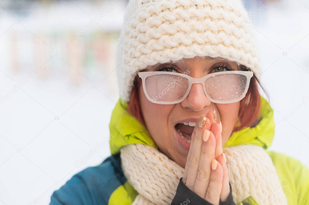 Portrait of a woman in glasses covered with hoarfrost. The girl is freezing and forgot gloves in very cold weather and blows on her bare hands