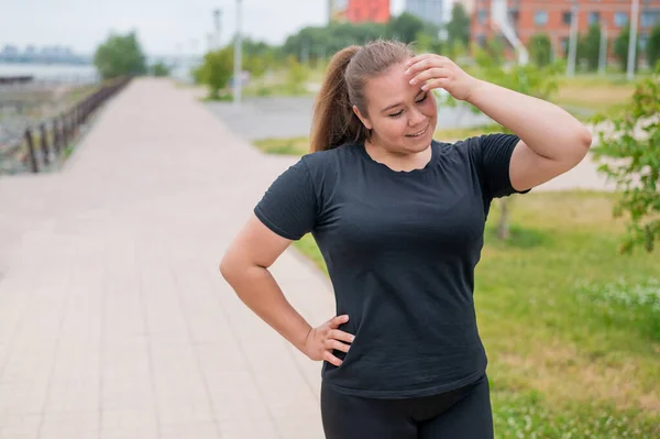 Eine übergewichtige junge Frau joggt im Freien. Dicke schöne lächelnde Mädchen in einem schwarzen Trainingsanzug ist in Fitness für die Gewichtsabnahme am Wasser engagiert. Eine Frau läuft an einem Sommertag. — Stockfoto