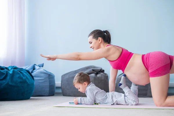 En gravid kvinna i sportkläder gör gymnastik medan hennes son spelar i mobilen. Flicka på yoga med sitt barn. Fitness mamma och barn. — Stockfoto