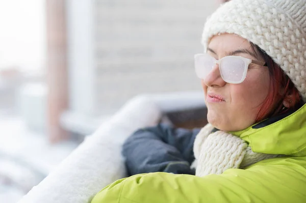 Μια δυσαρεστημένη λευκή γυναίκα στέκεται στο μπαλκόνι ενός πλίνθινου σπιτιού το χειμώνα, δείχνοντας τη γλώσσα και τον αντίχειρά της προς τα κάτω. Κορίτσι με γυαλιά καλυμμένα με πάγο στο δρόμο — Φωτογραφία Αρχείου