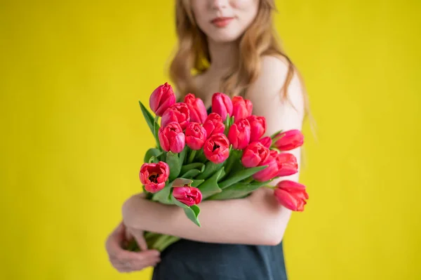 Eine unkenntliche Frau mit einem Strauß roter Tulpen auf gelbem Grund. Ein Mädchen in einem schwarzen Kleid hält einen Blumenstrauß in der Hand. Ein Geschenk zum Internationalen Frauentag. Frühjahrsferien. — Stockfoto
