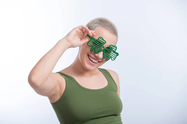 Portrait d'une jeune femme caucasienne avec une coupe de cheveux mans dans un T-shirt vert et des lunettes gaies sur un fond blanc. La fille célèbre st patricks jour — Photo