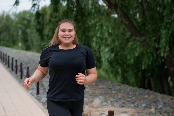 Een zwaarlijvige jonge vrouw die buiten jogt. Fat mooi lachend meisje in een zwart trainingspak is bezig met fitness voor gewichtsverlies aan het water. Een vrouw rent op een zomerdag. — Stockfoto