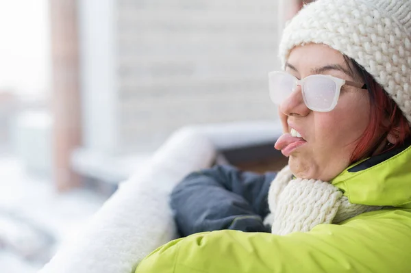 Uma mulher caucasiana descontente fica na varanda de uma casa de tijolos no inverno, mostrando a língua e o polegar para baixo. Menina em óculos cobertos de gelo na rua ao ar livre — Fotografia de Stock