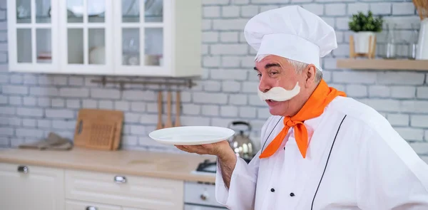 Un chef âgé sympathique tient une assiette dans la cuisine. Un homme aux cheveux gris avec une épaisse moustache grise travaille dans la cuisine — Photo