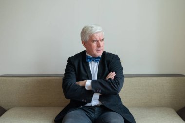 Memnun olmayan takım elbiseli ve papyonlu bir iş adamı kollarını göğsünde kavuşturarak oturur..