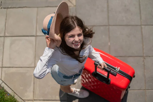 Top uitzicht op mooie jonge Kaukasische vrouw in de zomer hoed poseren met rode koffer buiten. Gelukkig lachend meisje gaat op vakantie met een grote bagage tas. Charmante dame in korte broek op een hete zonnige dag. — Stockfoto