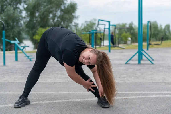 Dikke jonge vrouw doet stretching oefeningen buiten. Overgewicht meisje maakt bochten voor joggen. — Stockfoto