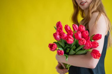 Sarı arka planda bir buket kırmızı lalesi olan tanınmayan bir kadın. Siyah elbiseli bir kız bir kucak dolusu çiçek tutuyor. Uluslararası Kadınlar Günü için bir hediye. Bahar tatili.