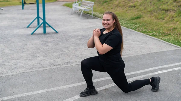 Krásná tlustá dívka v černé teplákové soupravě se zabývá fitness na sportovním hřišti. Mladá žena se vrhá ven za teplého letního dne. Zdravý životní styl a hubnutí. — Stock fotografie