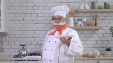 Dost canlısı yaşlı bir şef mutfakta bir tabak tutuyor. Gri saçlı, kalın gri bıyıklı bir adam mutfakta çalışıyor.