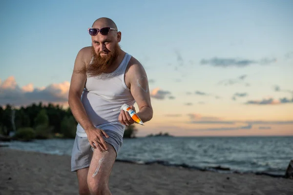 Uomo barbuto che applica la protezione solare sul corpo mentre in piedi sulla spiaggia. — Foto Stock