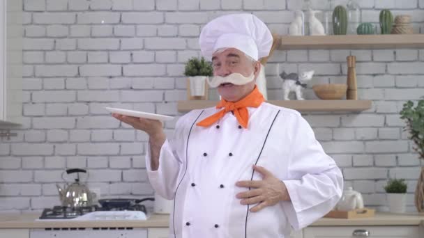En trevlig äldre kock håller en tallrik i köket. En gråhårig man med tjock grå mustasch jobbar i köket. — Stockvideo
