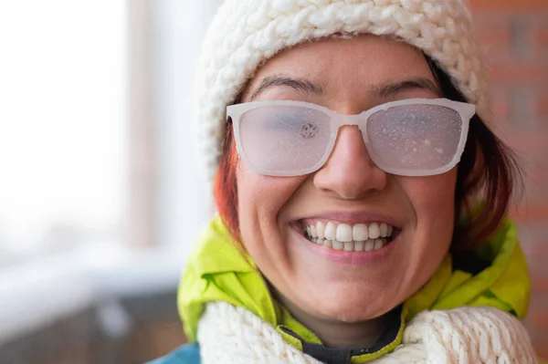 Den leende vita kvinnan står på balkongen i ett tegelhus på vintern. Glad flicka i istäckta glasögon på gatan utomhus — Stockfoto