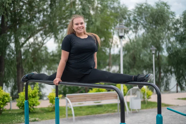Eine schöne lächelnde, übergewichtige junge Frau streckt sich auf unebenen Stufenbarren im Freien. Dicke Turnerinnen beim Fitnesstraining auf dem Sportplatz. Sport draußen an einem warmen Sommertag. — Stockfoto