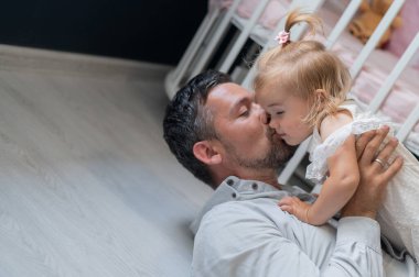 Kafkasyalı adam sırt üstü uzanıyor ve kızını çocuk odasında onun üstünde tutuyor.