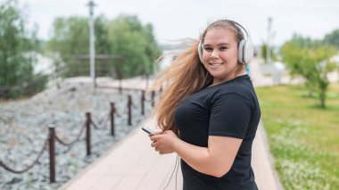 Kulaklıklı genç şişman kadın dışarıda yürüyor ve akıllı telefondan müzik dinliyor.