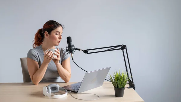 En kvinna sänder online på en bärbar dator och dricker kaffe. Kvinnlig radiovärd på arbetsplatsen — Stockfoto