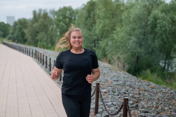 Eine übergewichtige junge Frau joggt im Freien. Dicke schöne lächelnde Mädchen in einem schwarzen Trainingsanzug ist in Fitness für die Gewichtsabnahme am Wasser engagiert. Eine Frau läuft an einem Sommertag. — Stockfoto