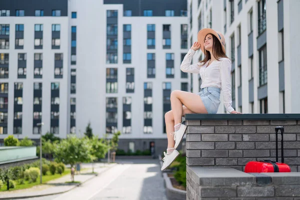 Hermosa joven en pantalones cortos y un sombrero posando mientras está sentada en un banco de piedra. Un retrato completo de una chica caucásica en un caluroso día de verano. —  Fotos de Stock