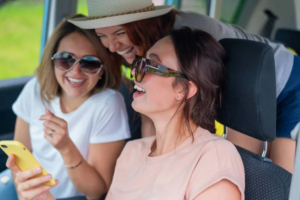 Três namoradas felizes fazem uma viagem. Mulheres Andar de carro, olhar para o telefone e rir. — Fotografia de Stock