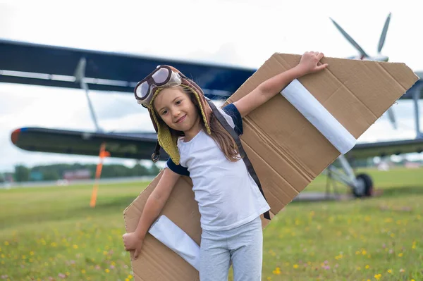 Ein kleines Mädchen spielt einen Piloten auf dem Hintergrund eines kleinen Flugzeugs mit Propeller. Ein Kind im Anzug mit Pappflügeln träumt vom Fliegen — Stockfoto