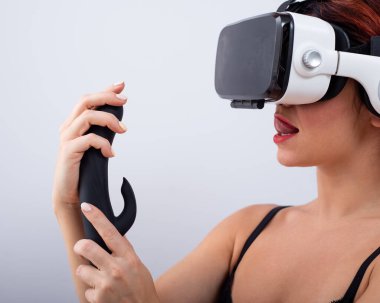 Caucasian red-haired woman wearing virtual reality glasses holding black dildo and looking lustfully.