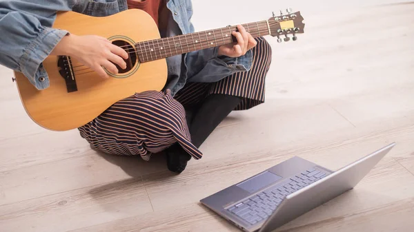 Junge Hipsterfrau sitzt zu Hause auf dem Fußboden und schaut sich am Laptop Gitarren-Tutorials an. Musik im Fernstudium in Quarantäne — Stockfoto