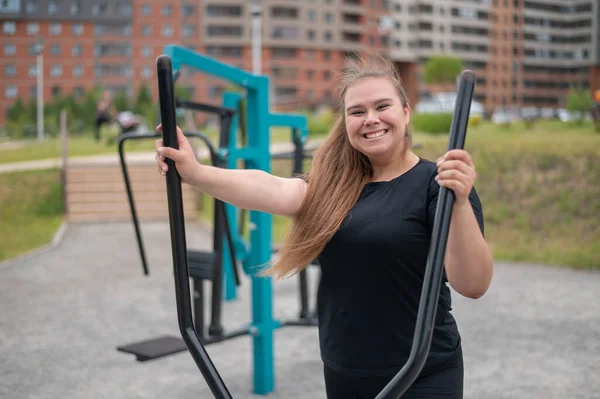 Schöne lächelnde übergewichtige junge Frau macht Fitness auf einem Ellipsoid-Simulator im Freien. Dicke Mädchen trainieren auf dem Sportplatz zum Abnehmen. Sport draußen an einem warmen Sommertag. — Stockfoto