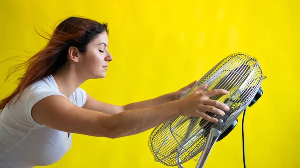 Krásná rusovlasá žena stojí nad velkým elektrickým ventilátorem na žlutém pozadí. Dívka s vlasy rozvíjejícími se ve větru. Zařízení pro chlazení vzduchu. — Stock fotografie