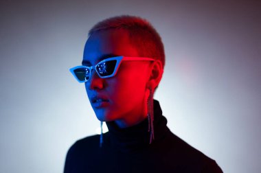 Stüdyoda kısa erkek saç kesimli, güneş gözlüklü, neon ışıklı genç bir kadının portresi.