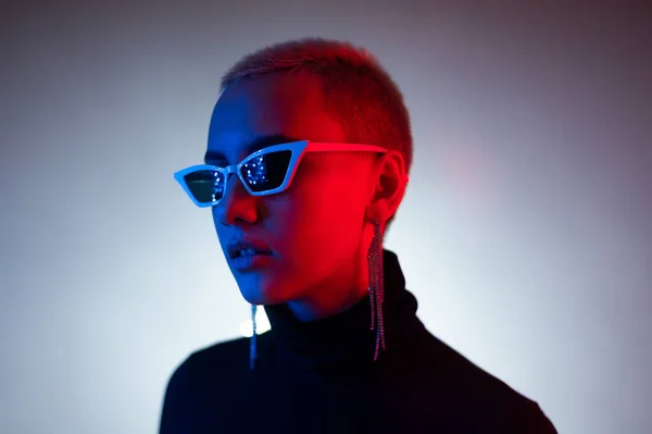 Porträtt av en ung kvinna med en kort manlig frisyr i solglasögon i neonljus i studion — Stockfoto
