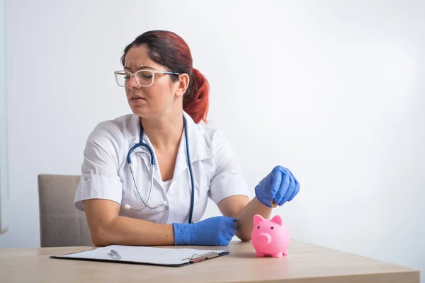 Rozrušený ženský doktor dává minci do prasečí banky na bílém pozadí. — Stock fotografie