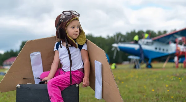 Ein kleines Mädchen im Pilotenkostüm mit Pappflügeln rennt vor der Kulisse des Flugzeugs über den Rasen. Ein Kind mit Hut und Brille träumt davon, im Flugzeug zu fliegen. — Stockfoto