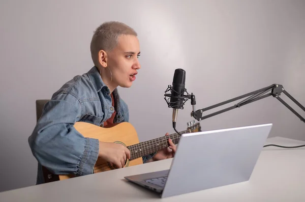 Junge Hipsterfrau nimmt im heimischen Tonstudio einen Song auf. Ein Mädchen spielt eine Akustikgitarre singt in ein Mikrofon und nimmt Video auf einem Laptop auf — Stockfoto