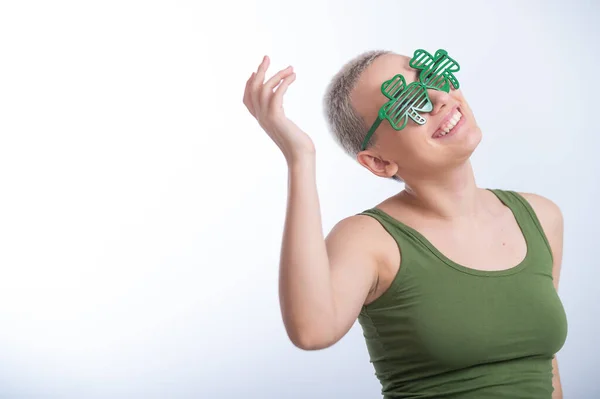 Портрет молодой белой женщины с мужской стрижкой в зеленой футболке и веселыми очками на белом фоне. Девушка празднует День Святого Патрика — стоковое фото