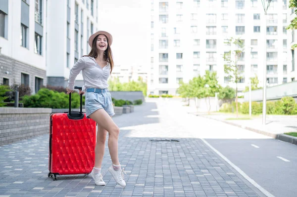 Jonge blanke vrouw in een hoed en korte broek staat op straat met een rode koffer. — Stockfoto